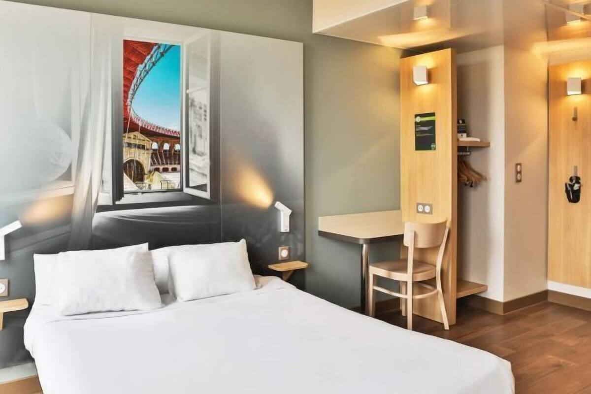 A hotel room with a bed, a desk, a chair, a wall-mounted TV, and a door with a coat rack.