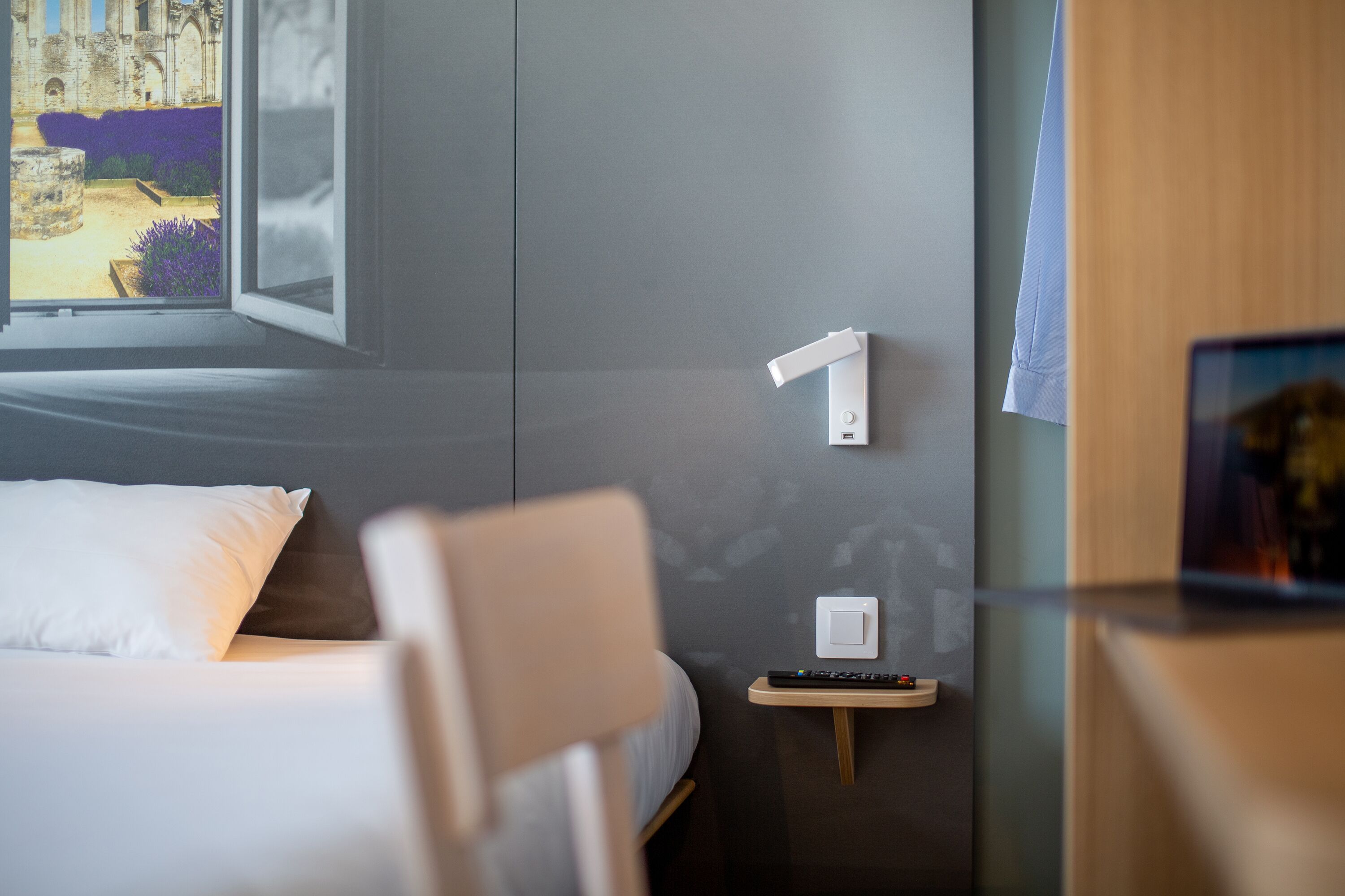A hotel room with a bed, a chair, a bedside table with a remote control, and a wall-mounted light switch.