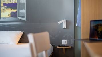 A hotel room with a bed, a chair, a bedside table with a remote control, and a wall-mounted light switch.