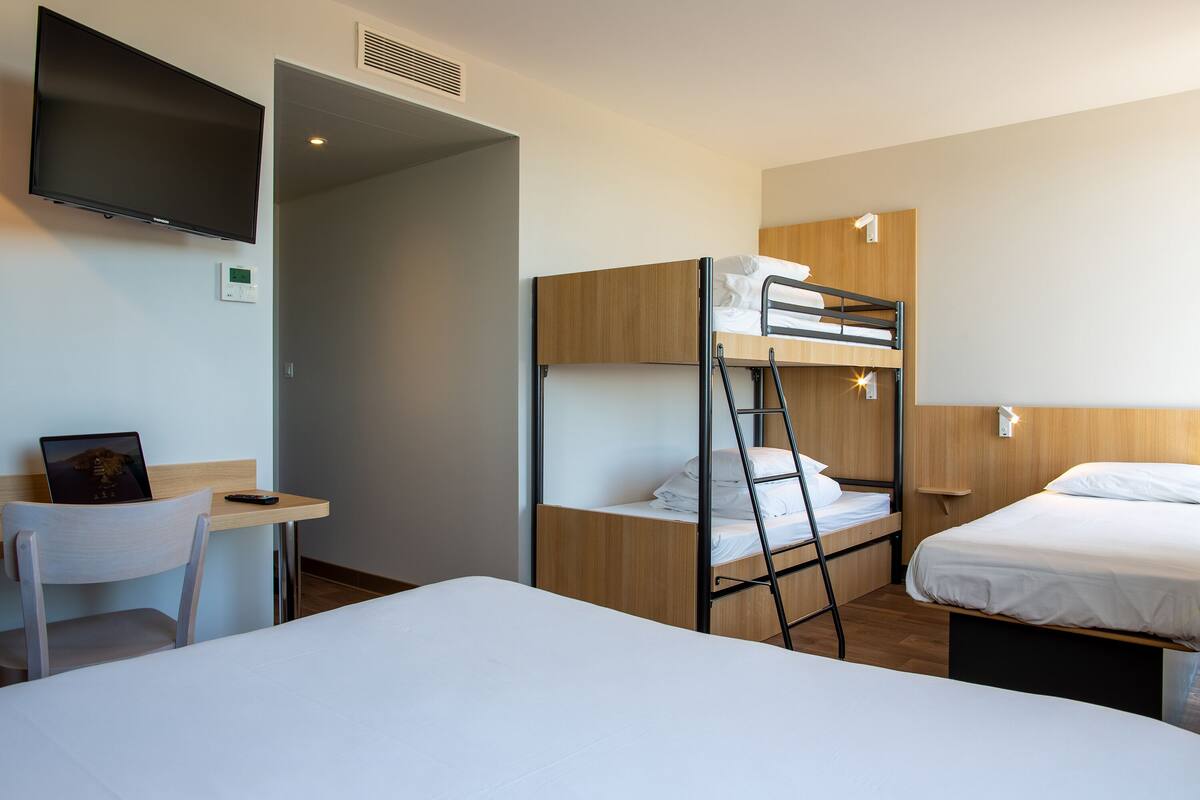 A hotel room with bunk beds, a desk with a chair, a TV mounted on the wall, and a bedside table.