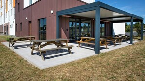A modern building with outdoor picnic tables and a covered seating area. - B&B HOTEL LES HERBIERS (Les Herbiers)