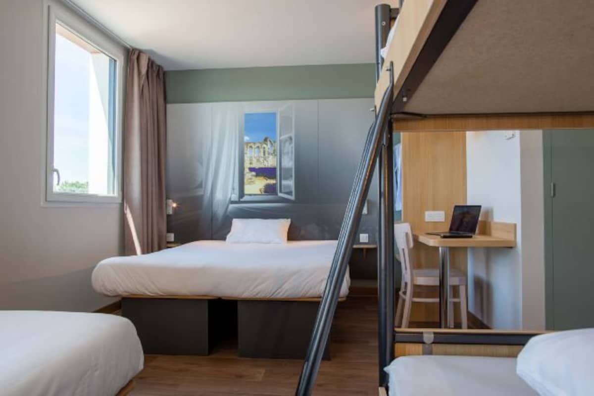 Una habitación con dos camas, un escritorio y vistas a un edificio a través de una ventana.