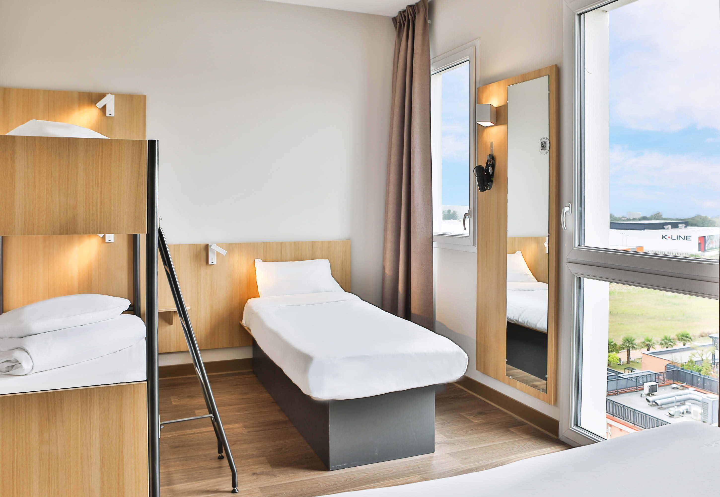 A modern hotel room with a bed, a bedside table, a mirror, and a window with a view of the city.