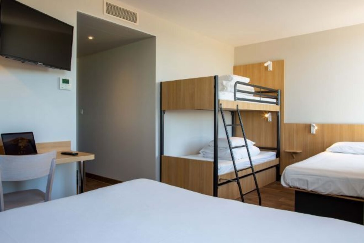 Habitación de hotel con literas, un escritorio, una silla, un televisor montado en la pared y una ventana pequeña.