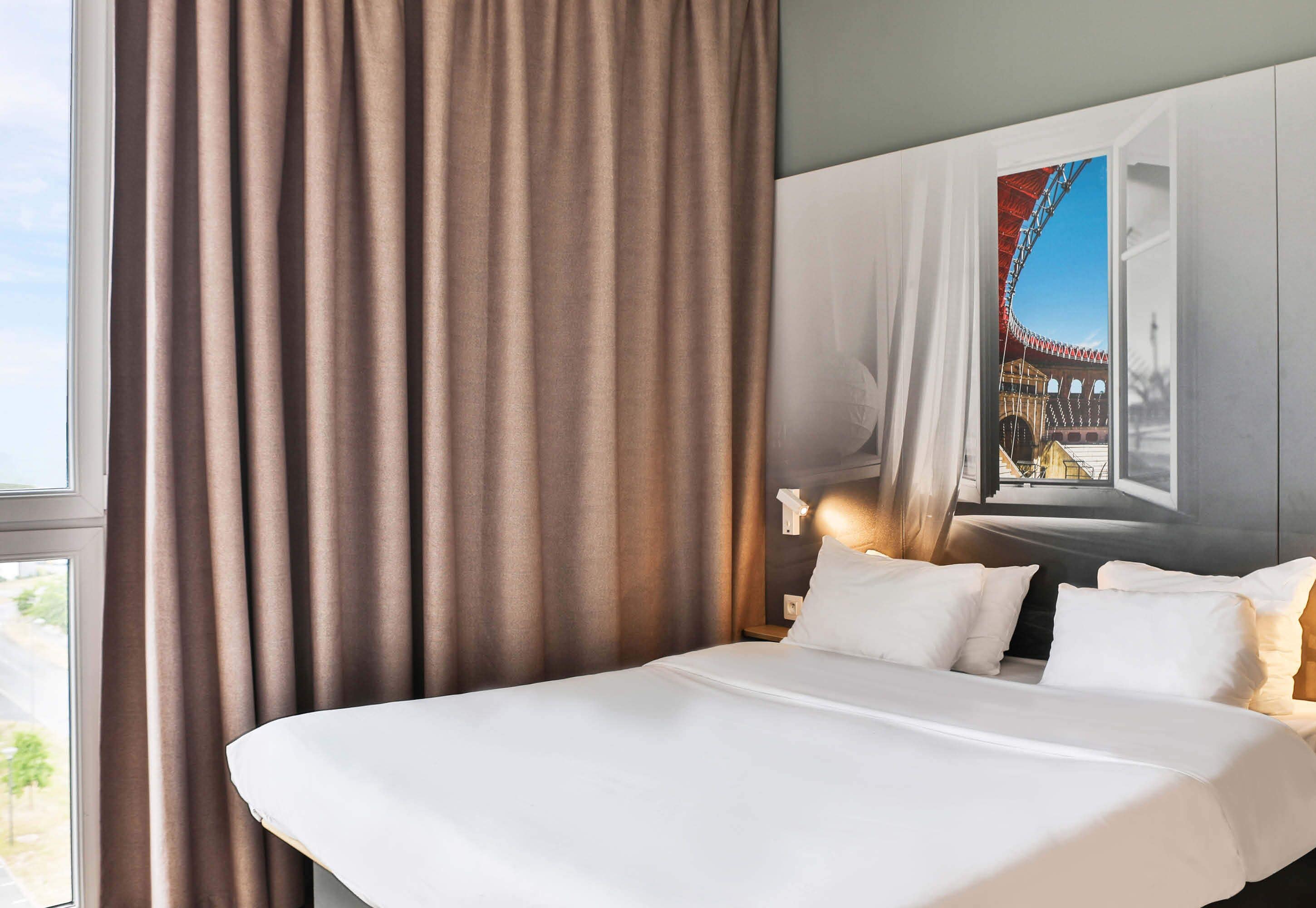 A hotel room with a large bed, a window with curtains, a wall-mounted lamp, and a large window with a view of a building.