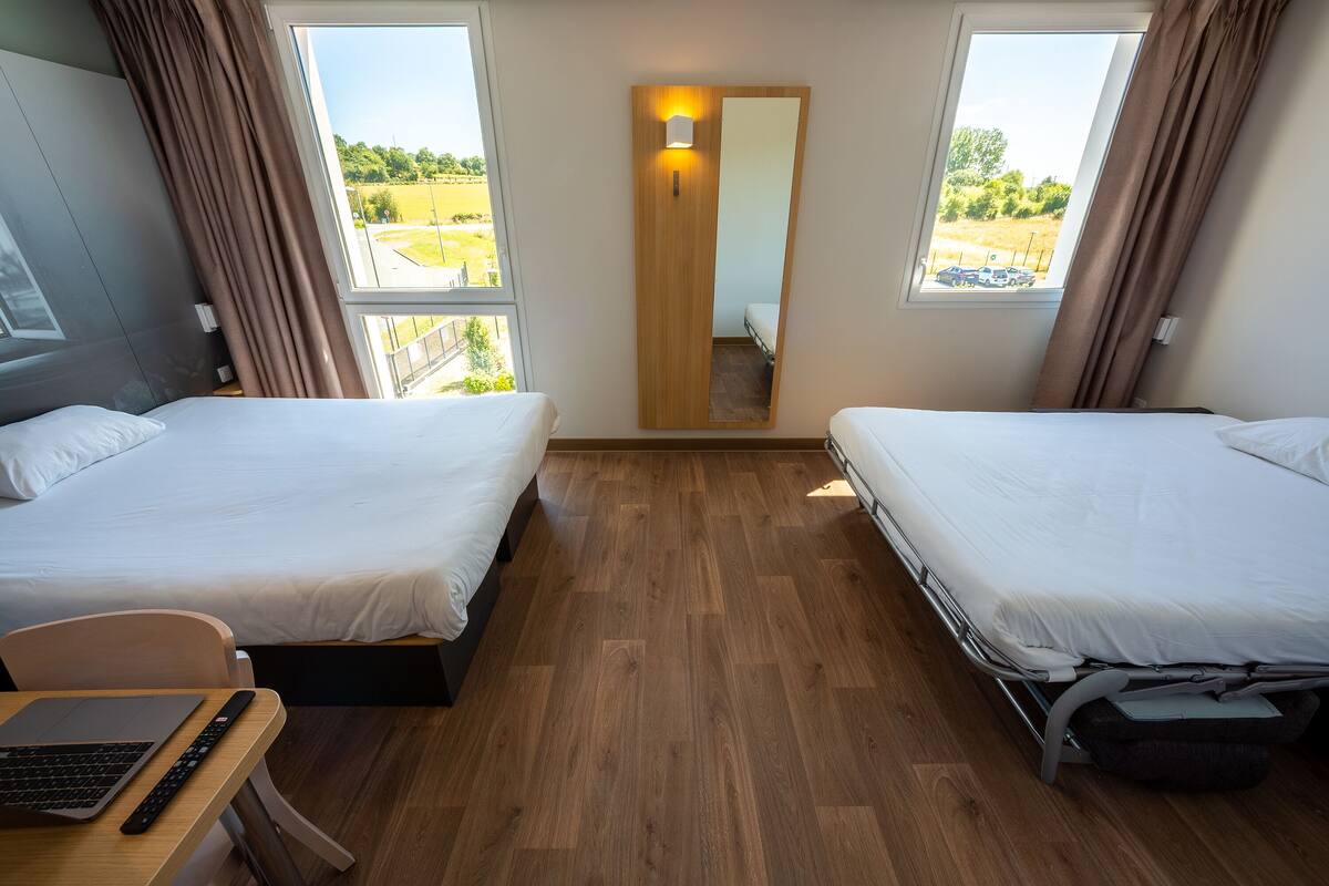 A hotel room with two beds, a desk with a laptop, and a large window with a view of a field.