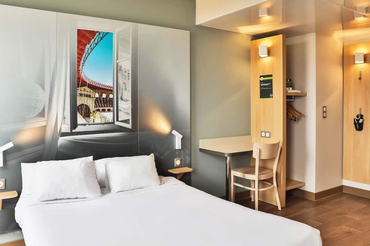A hotel room with a bed, a desk, a chair, a wardrobe, and a window with a view of a building.