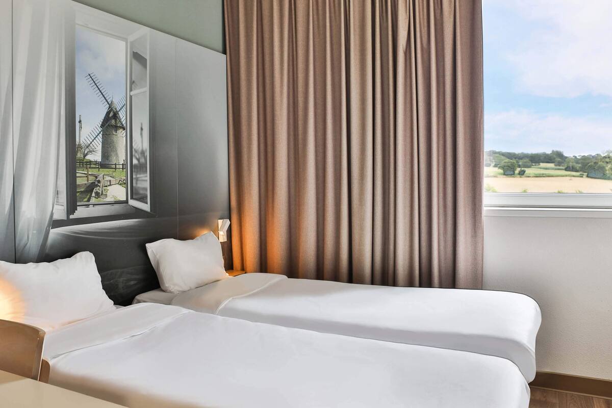 A hotel room with two beds, a large window with a view of a windmill, and a painting on the wall.