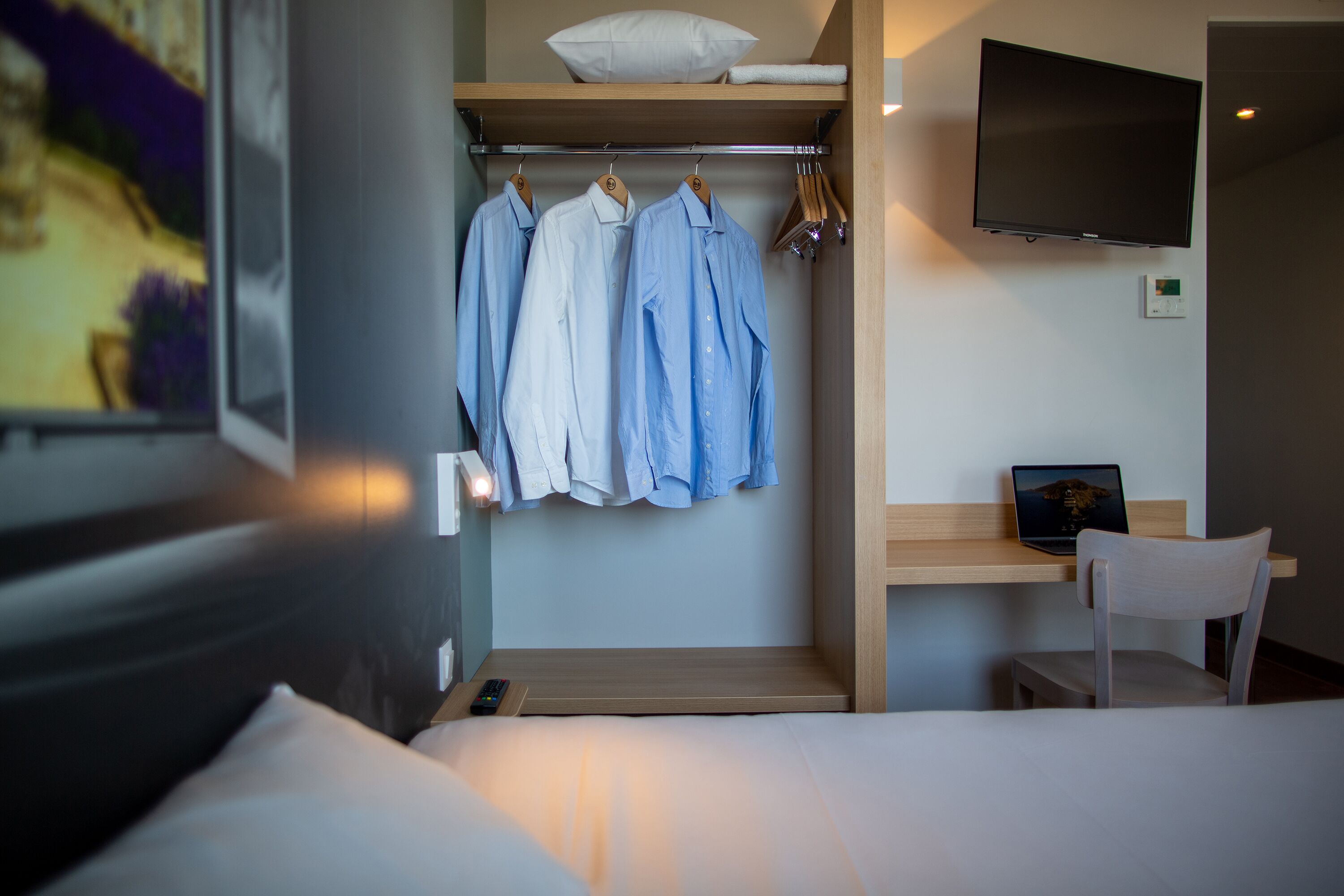 Habitación de hotel con cama, televisor montado en la pared, escritorio con una computadora portátil y perchero con ropa.