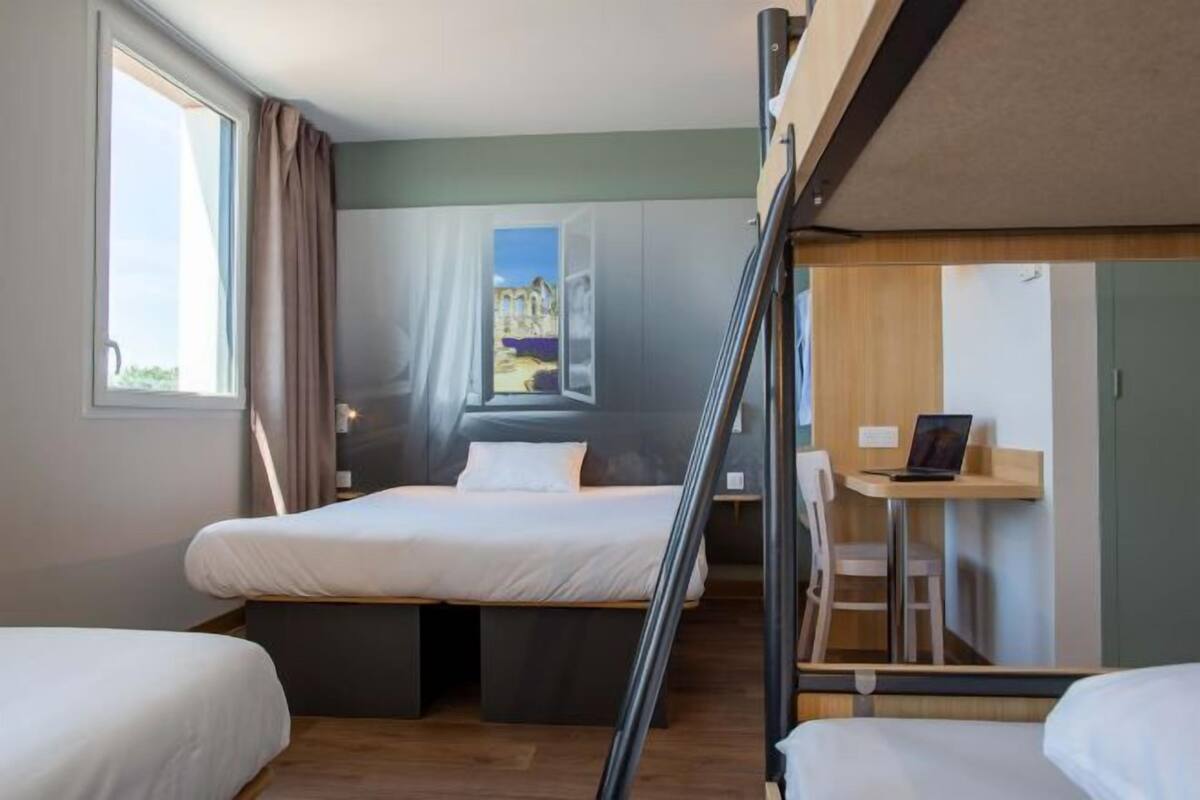A room with two beds, a desk, and a view of a building through a window.