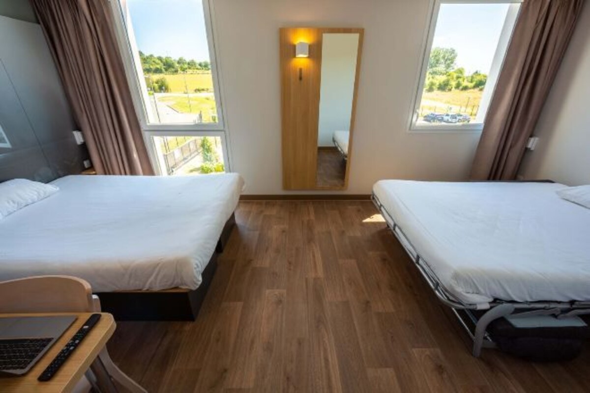 Habitación de hotel con dos camas, un escritorio con una computadora portátil, un espejo y vista al exterior.