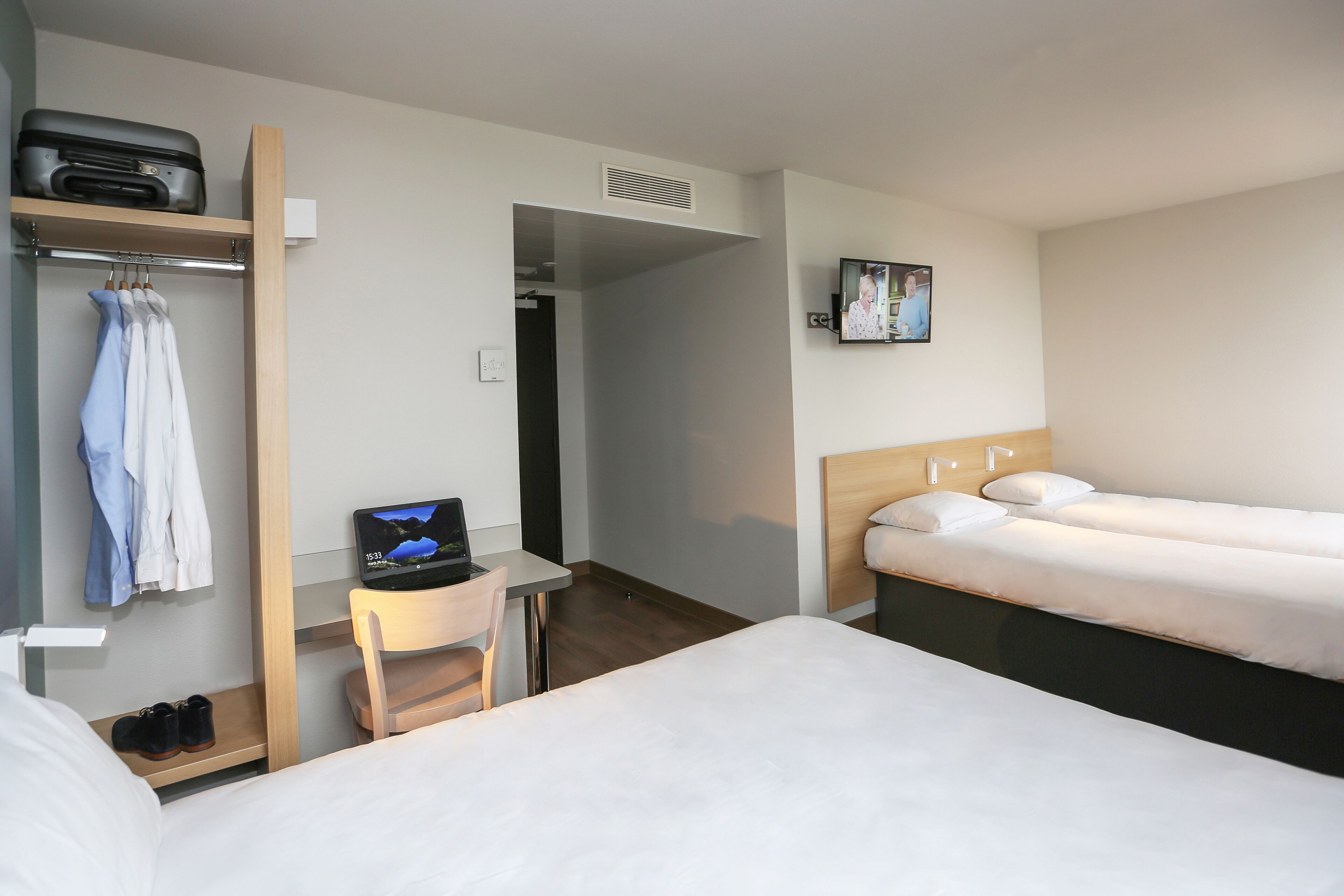 Une chambre d’hôtel équipée d’un lit, d’un bureau avec un ordinateur, d’une chaise, d’un porte-manteau et d’une télévision.