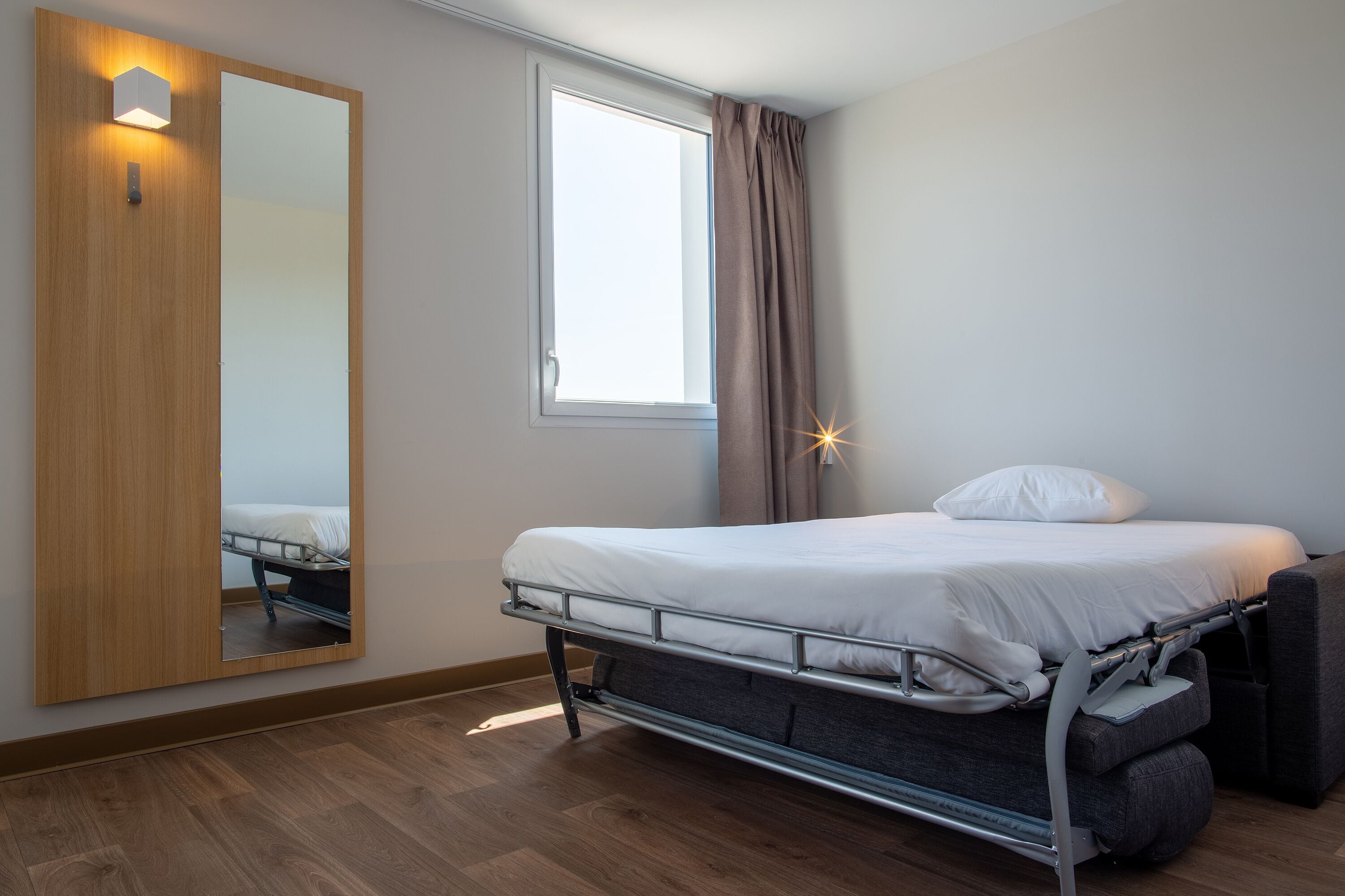 Una habitación pequeña con una cama, un espejo grande, una ventana con cortinas y suelo de madera.