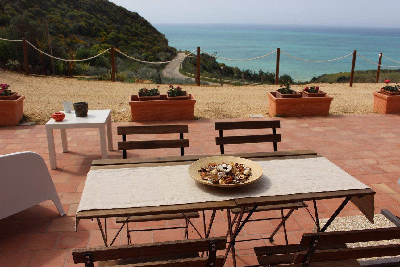 An outdoor seating area with a table, benches, and potted plants, overlooking a beach and ocean.