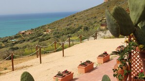 Un paysage côtier avec un sentier sablonneux, des cactus, des jardinières en terre cuite ornées de fleurs et, au loin, une tour perchée sur une colline.