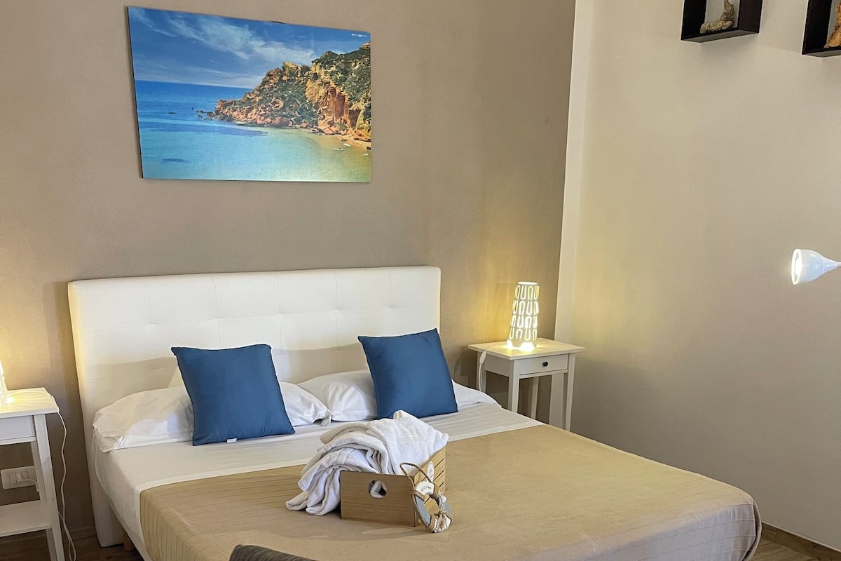 Una cama bien hecha con almohadas azules, una mesita de noche con lámpara y un cuadro de una escena costera en la pared.