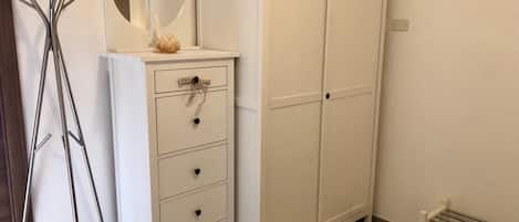 A white wardrobe with a mirror and a small decorative item on top, a white dresser with drawers, and a metal rack.