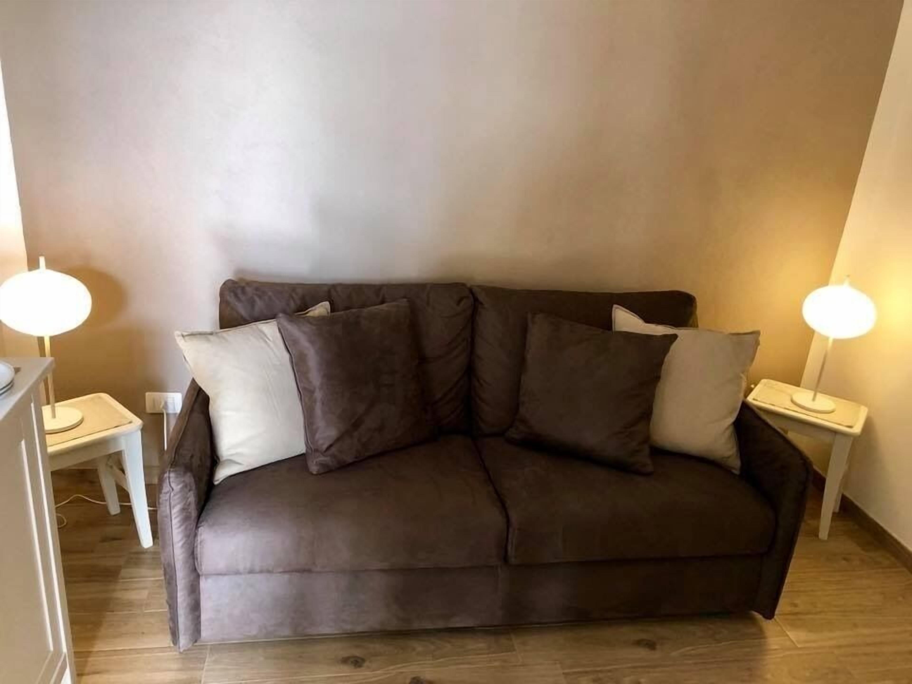 A brown sofa with multiple cushions, two bedside tables with lamps, and a wooden floor.