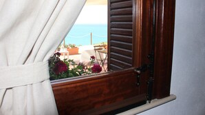 Une fenêtre avec des volets en bois, un rideau et une vue sur la mer, ainsi qu’un espace extérieur aménagé pour s’asseoir.