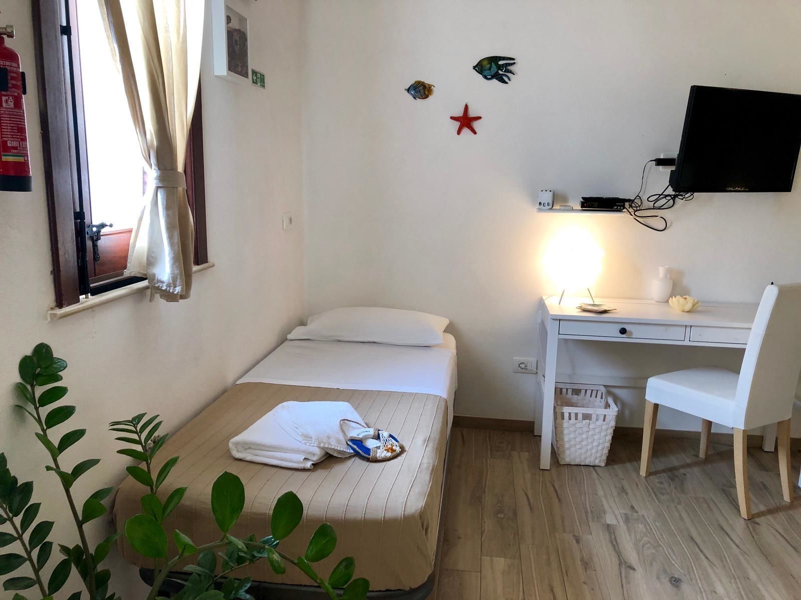 Ein kleiner, ordentlicher Raum mit einem Bett, einem Schreibtisch, einem Stuhl, einem Fernseher und einem Fenster mit Vorhängen.