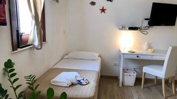 Ein kleiner, ordentlicher Raum mit einem Bett, einem Schreibtisch, einem Stuhl, einem Fernseher und einem Fenster mit Vorhängen.