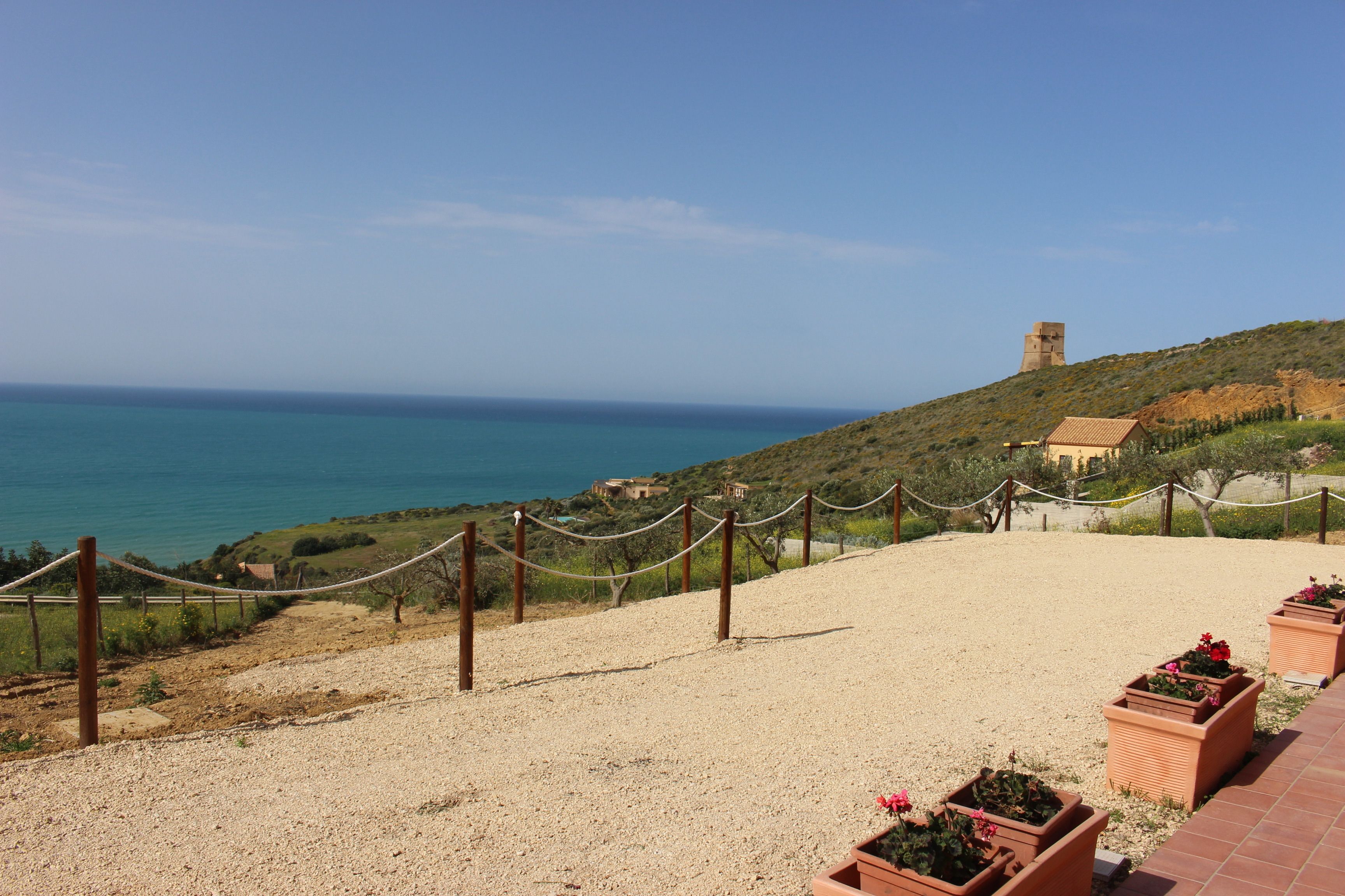 A coastal landscape with a clear view of the sea, a hill with a tower, and a fenced area with potted plants.