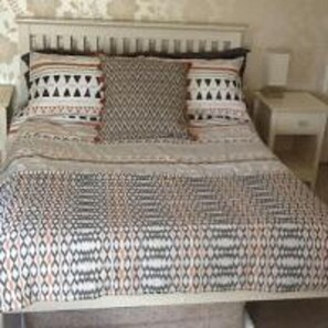 Una cama con colcha y almohadas estampadas, un cabecero de madera y una mesita de noche con una lámpara.