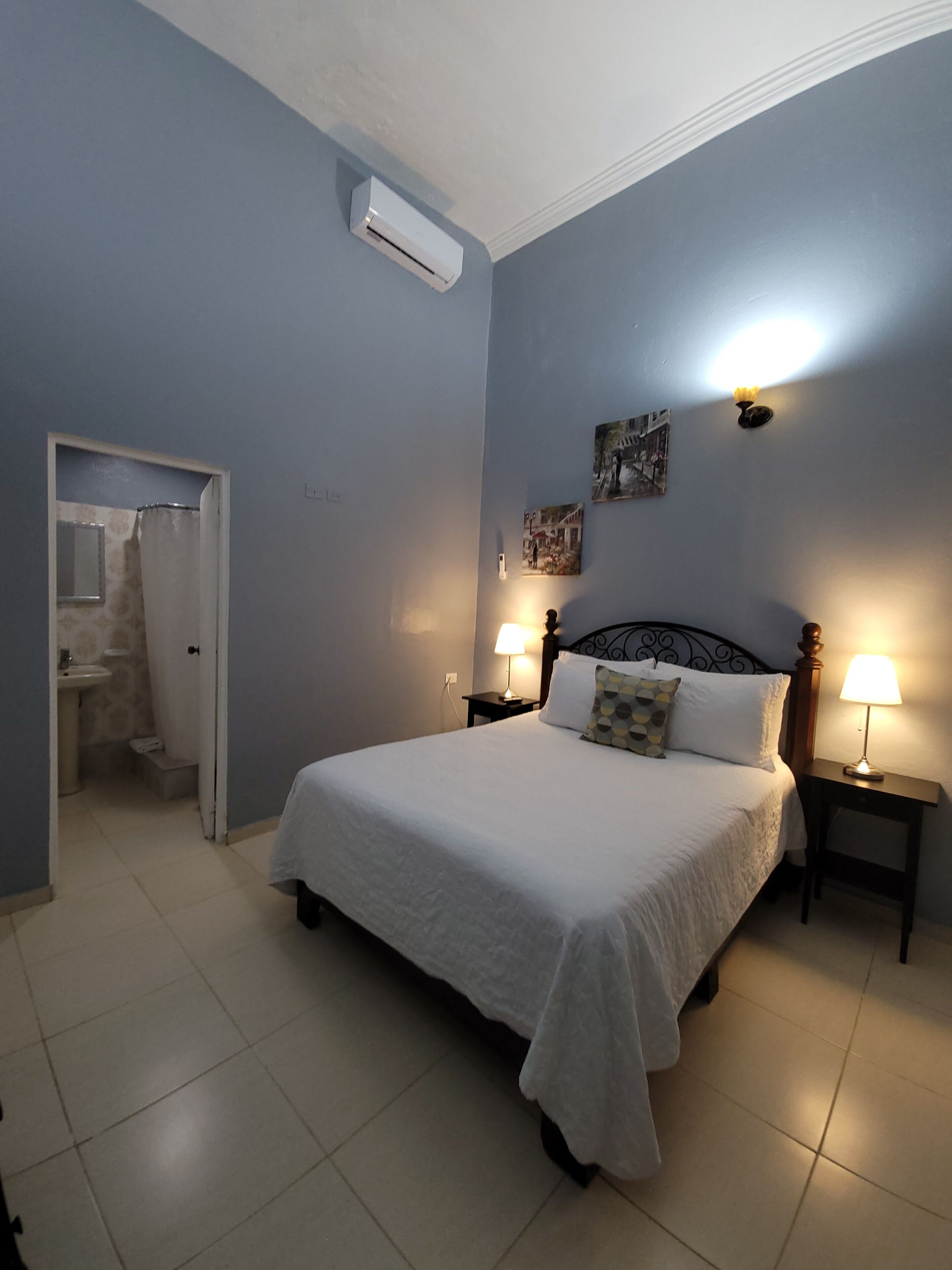 A bedroom with a bed, two bedside tables with lamps, an air conditioning unit, a door leading to a bathroom, and a wall with two pictures.