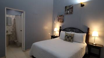 A bedroom with a bed, two bedside tables with lamps, an air conditioning unit, a door leading to a bathroom, and a wall with two pictures.