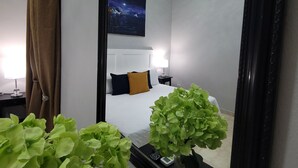 A hotel room with a large mirror, a bed with white bedding, a night sky painting on the wall, and a potted plant in the foreground. - Hotel Class Colonial (Santo Domingo)