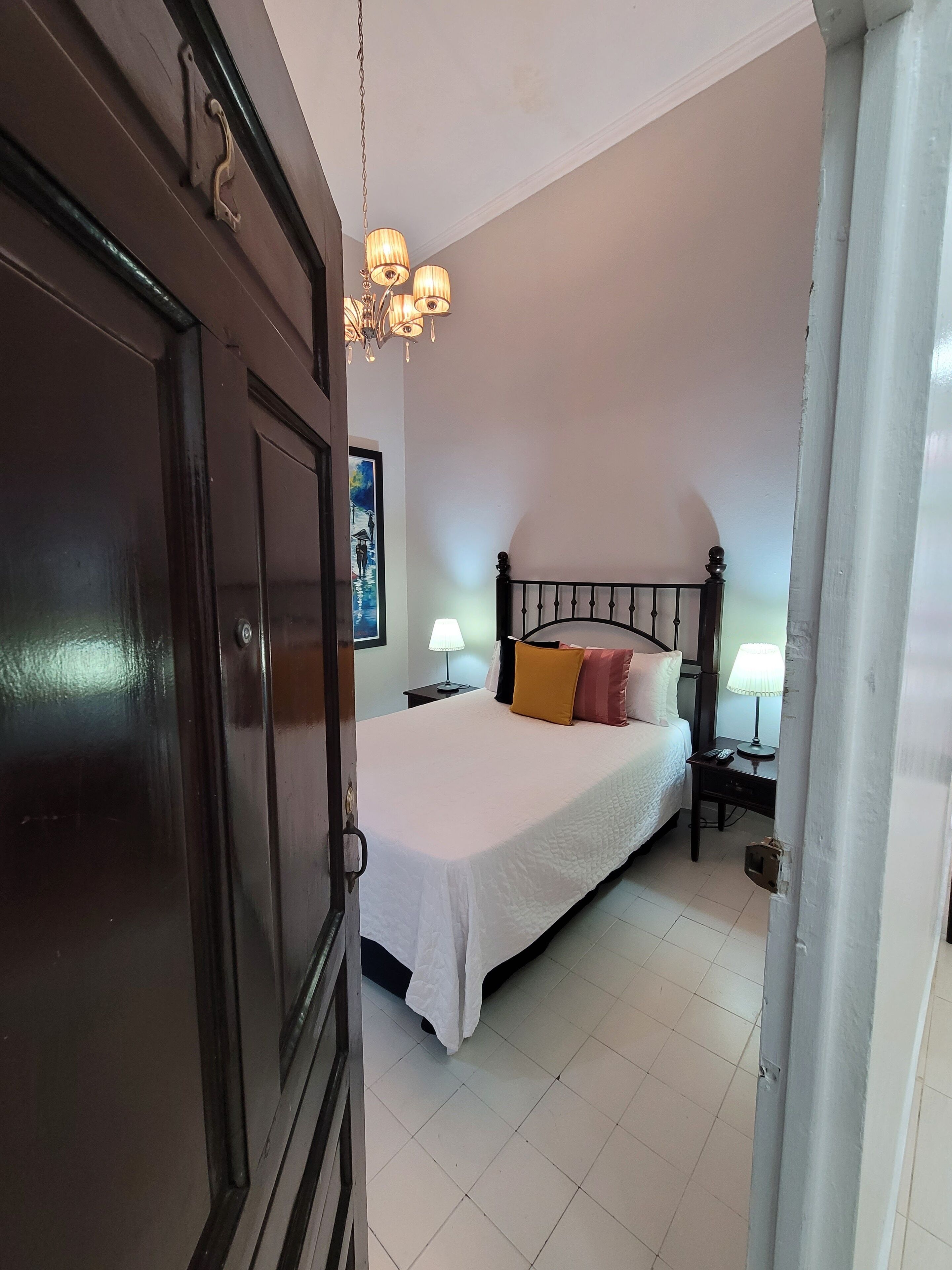 A bedroom with a bed, bedside lamps, a chandelier, and a door.