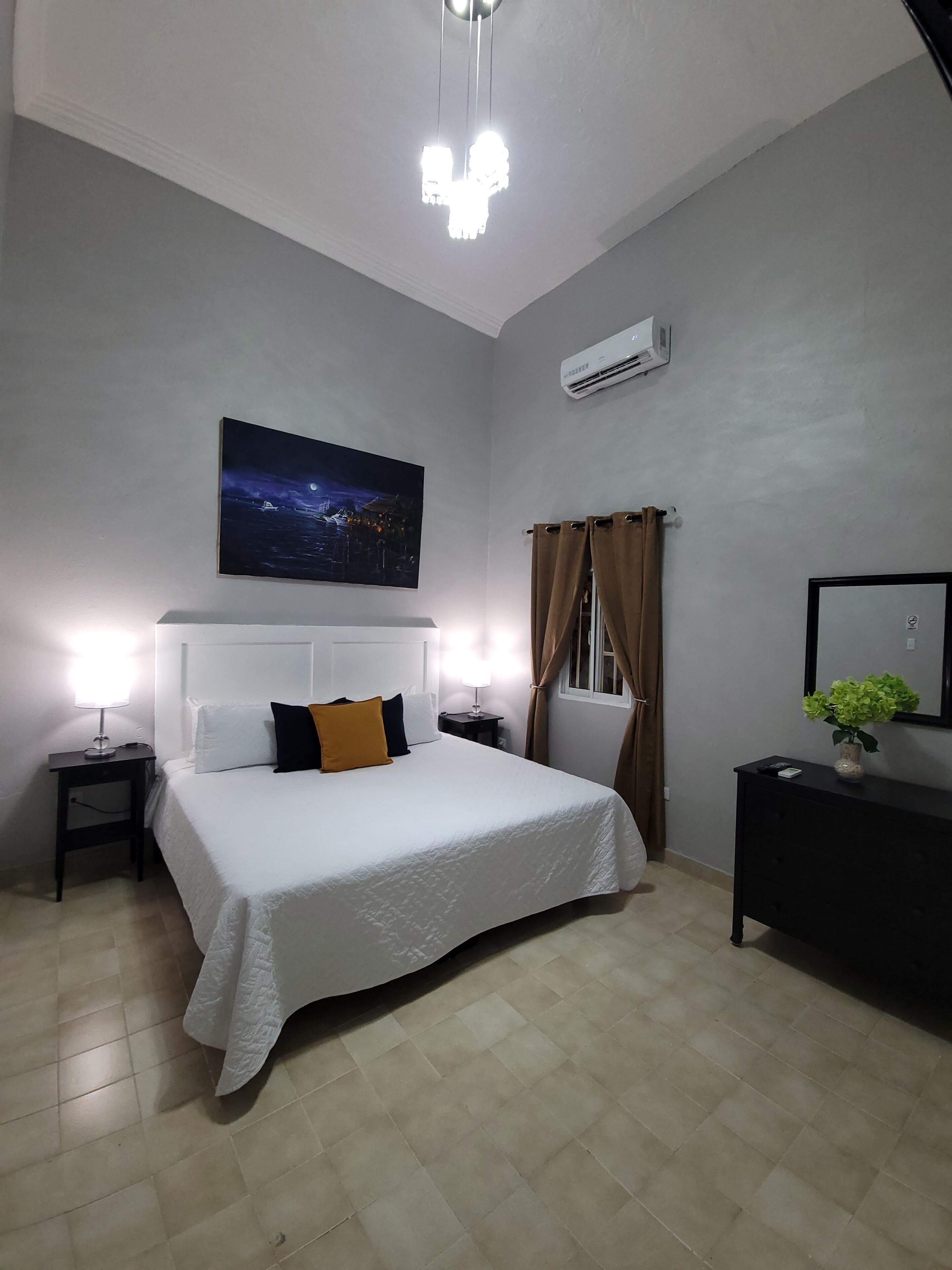 A bedroom with a large bed, a nightstand, a bedside lamp, a wall-mounted air conditioner, a window with curtains, and a nightstand with a plant.