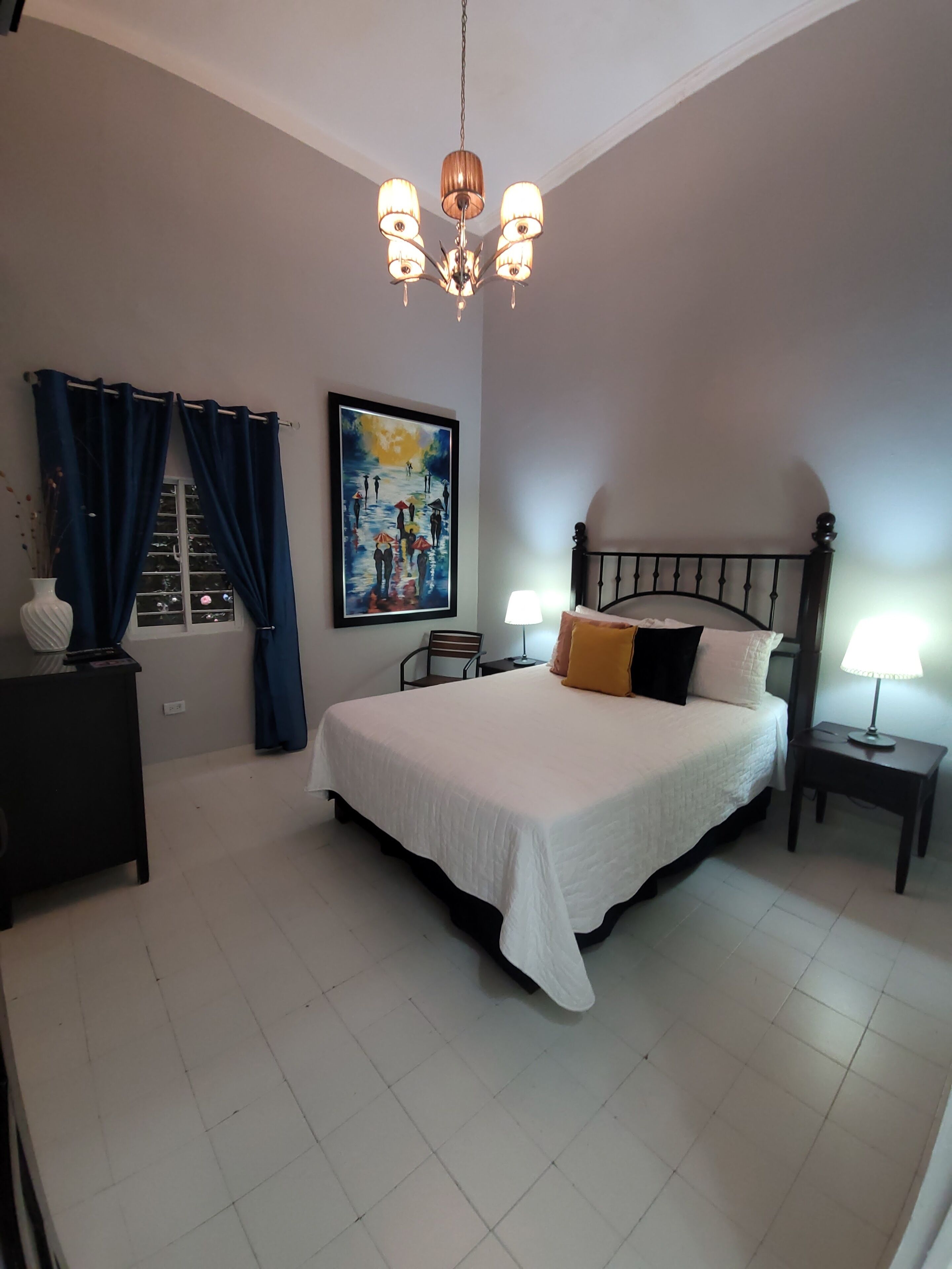 A bedroom with a large bed, two bedside tables with lamps, a chandelier, a painting on the wall, and a window with curtains.