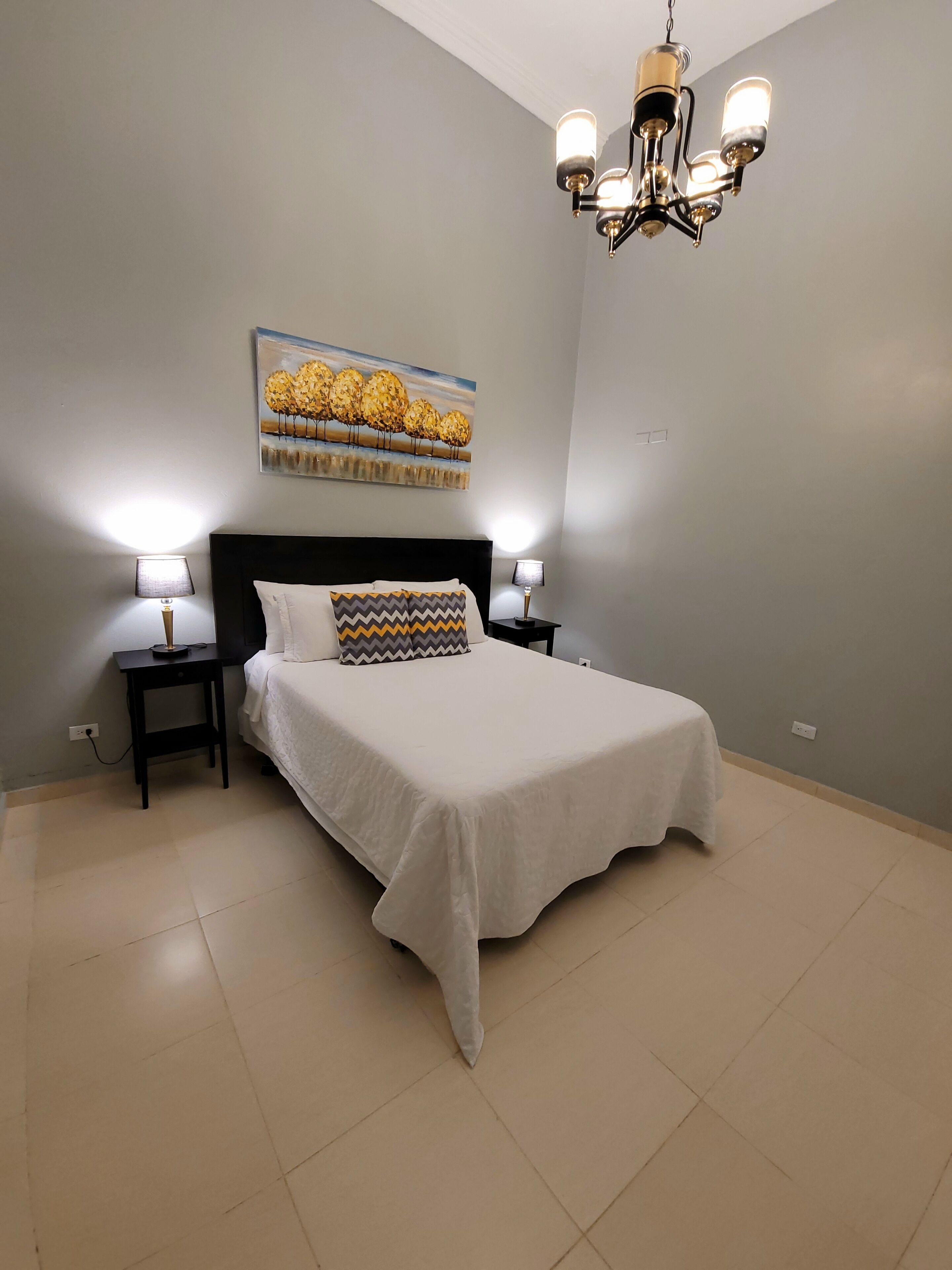 A bedroom with a bed, two bedside tables, a wall-mounted lamp, and a ceiling light fixture.