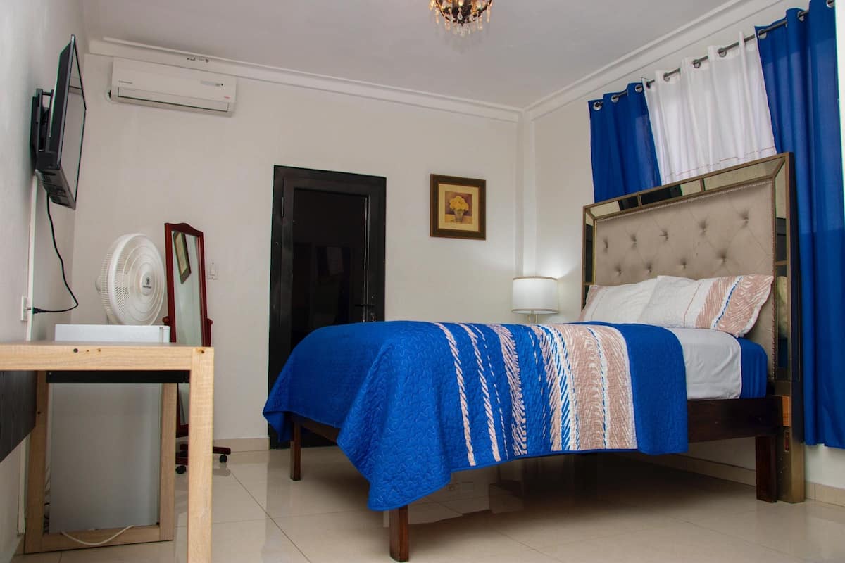 A bedroom with a bed, a desk with a computer, a fan, and a TV.