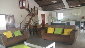 A spacious living area with two sofas, a coffee table, and a dining table with chairs. A staircase is visible in the background leading to an upper level. - The Sanctuary (Mahavelona)