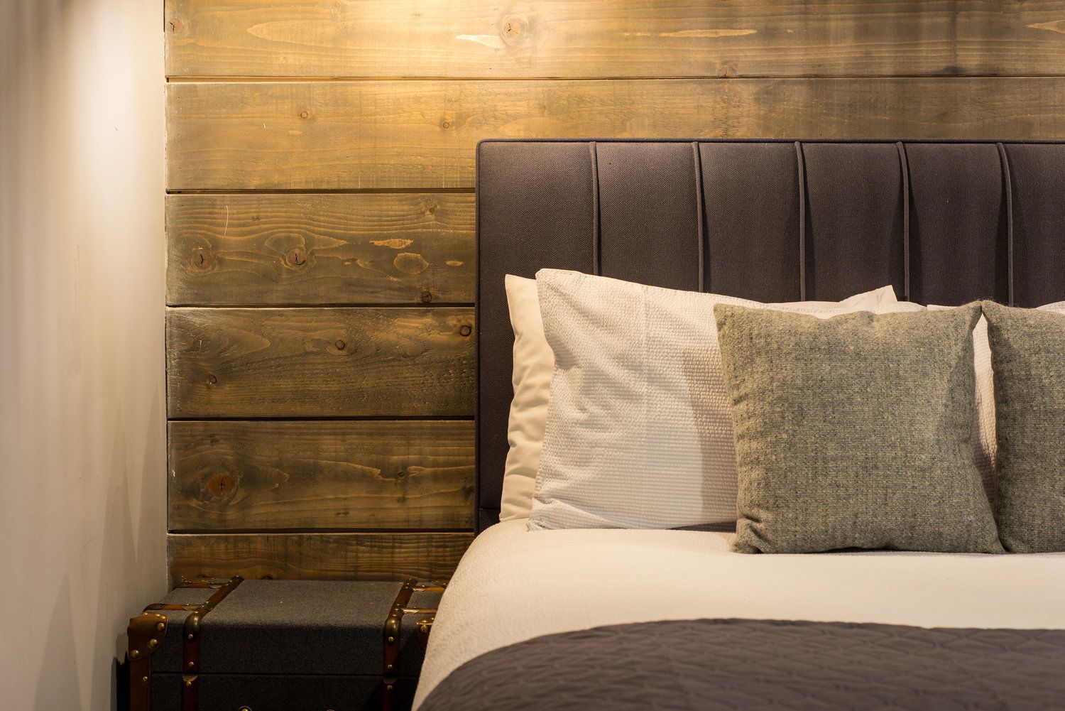 A bed with a dark headboard and multiple pillows, a small wooden nightstand, and a suitcase.