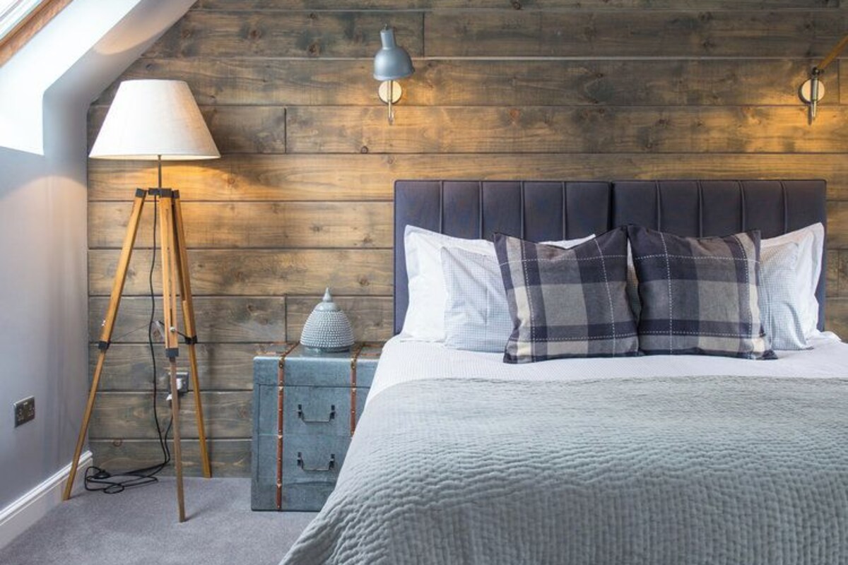 A bedroom with a sloped ceiling, a wooden headboard, a bed with white and plaid bedding, a floor lamp, and a wall-mounted light fixture.