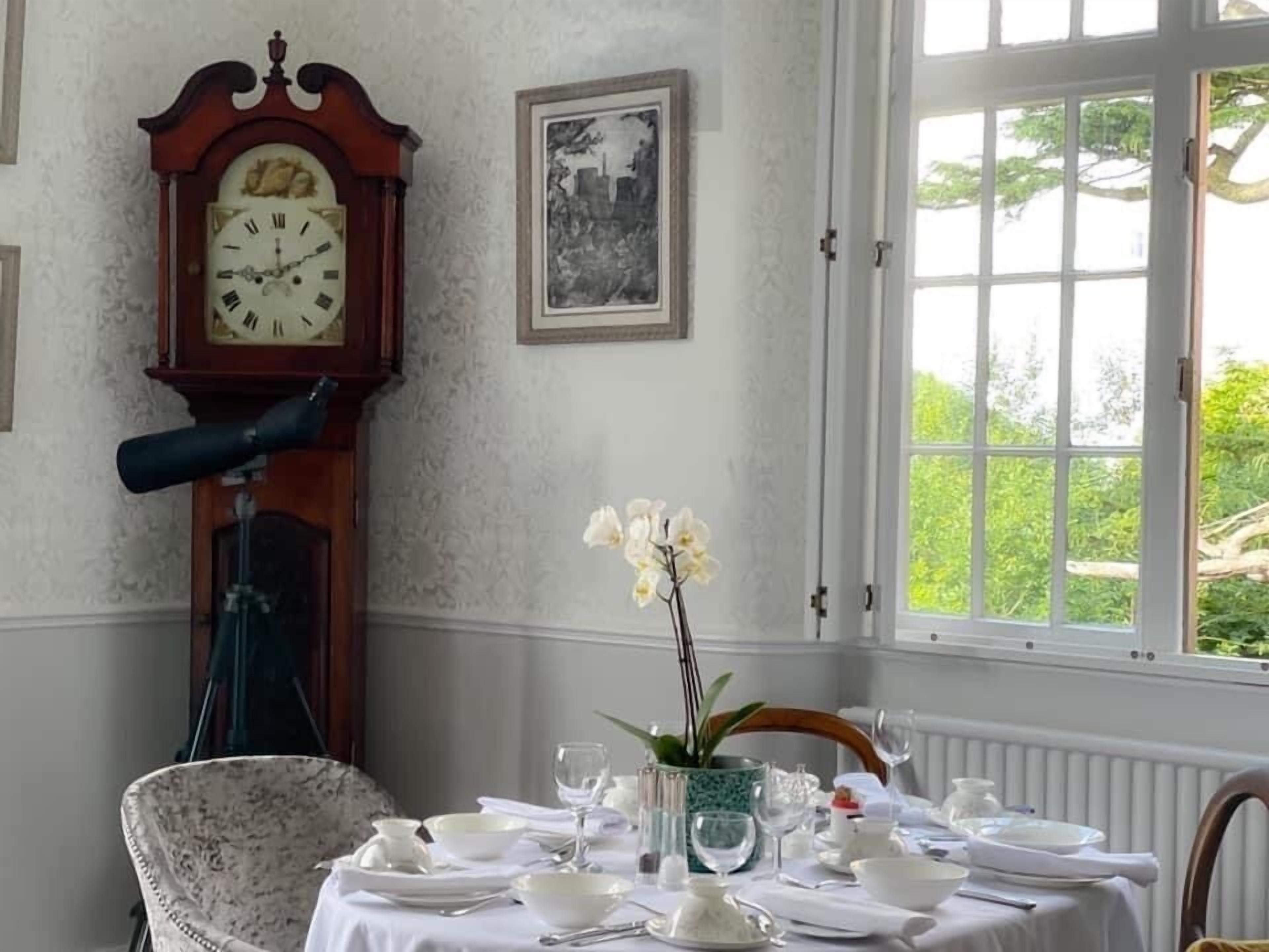 Comedor con una mesa dispuesta para cuatro, un reloj de pie, un cuadro enmarcado y un ventanal con vistas a follaje.