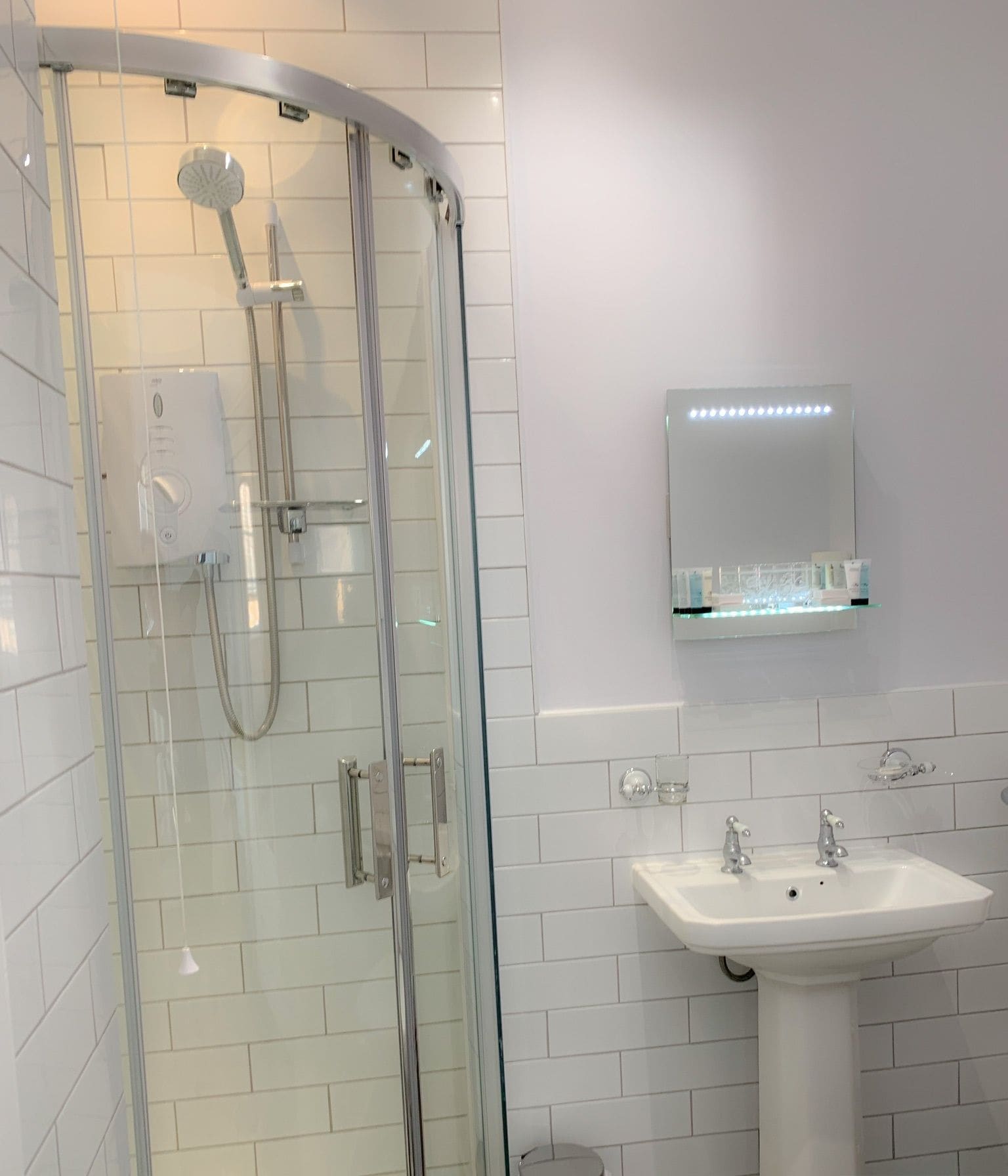 Un baño moderno con ducha envuelta en cristal, lavamanos pedestal y espejo con luces LED.