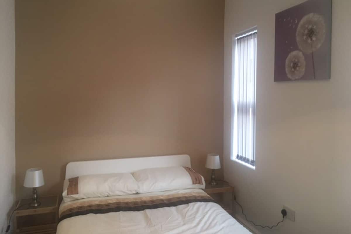 Een slaapkamer met een bed, nachtlampjes, een raam met zonwering en een kunstwerk aan de muur.