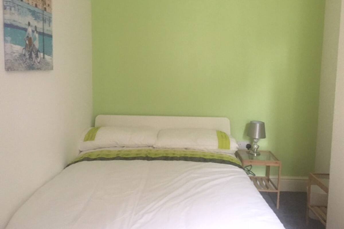 Een bed met witte beddengoed en een kussen met groene en witte strepen, een nachtkastje met een lamp, en een schilderij van een stadsbeeld aan de muur.