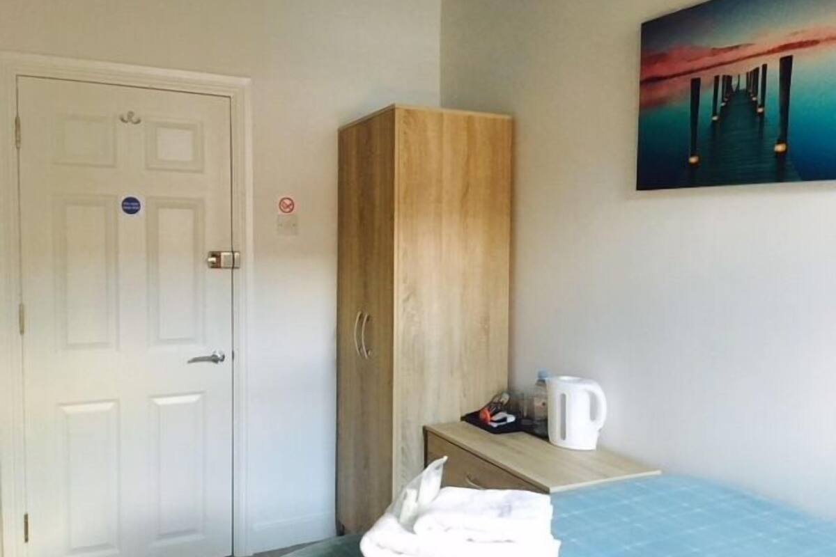 Habitación de hotel con una cama, un armario de madera, un cuadro de una puesta de sol y una tetera sobre una mesita.