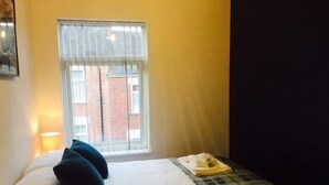Habitación de hotel con cama, ventana con persianas, lámpara y vista a un edificio de ladrillo.