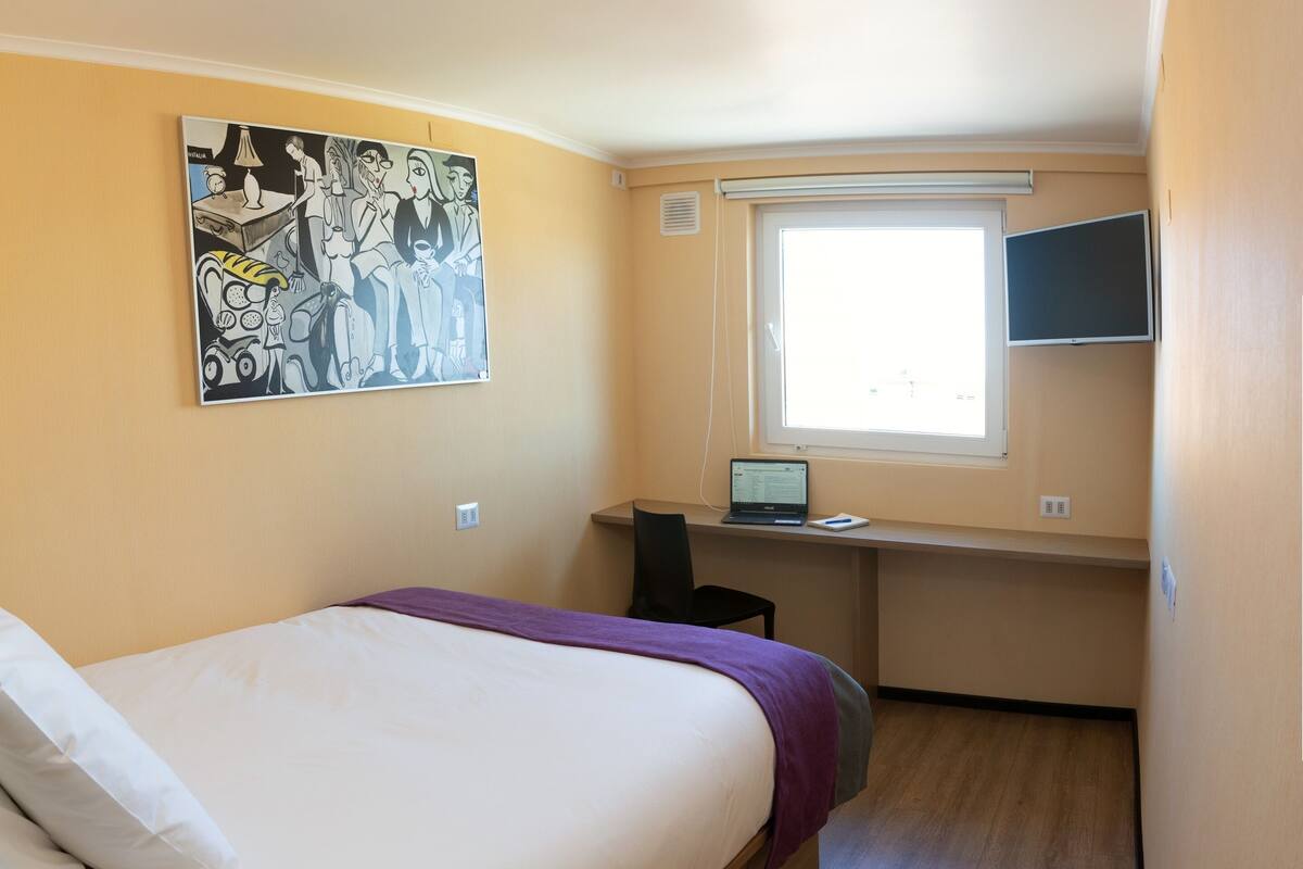 Un dormitorio con cama, un escritorio con ordenador portátil, una ventana y un cuadro en la pared.
