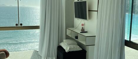 A hotel room with a bed, a bedside table, a TV, and a view of the sea.