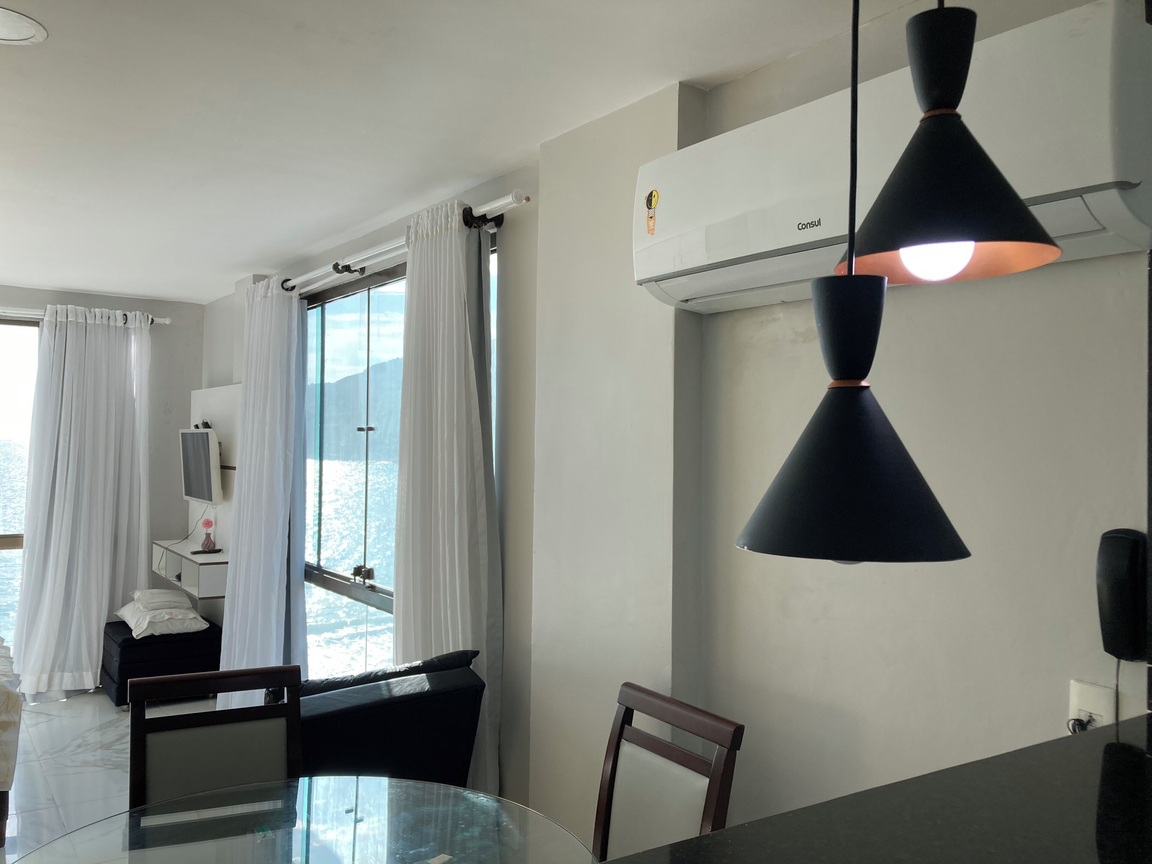 Quarto de hotel moderno com uma mesa de jantar de vidro, uma cadeira preta, uma luminária pendente preta, um ar condicionado branco e vista da cidade através de uma grande janela com cortinas brancas.