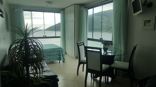 A room with a dining table, chairs, a potted plant, a bed with a green cover, and large windows with curtains. - Loft da Prainha (Arraial do Cabo)