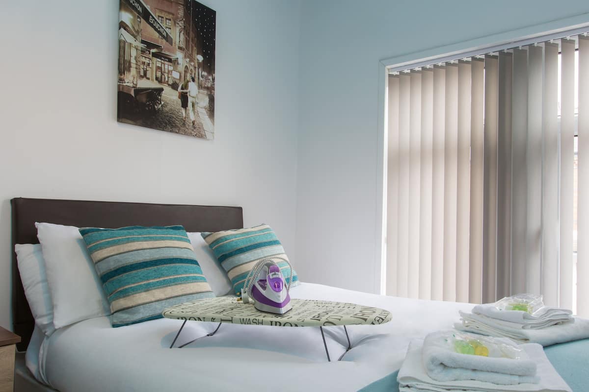 A neatly made bed with striped pillows, a small ironing board with an iron, and neatly folded towels.