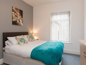A neatly made bed with a teal throw blanket and floral pillows, a nightstand with a lamp, and a window with blinds. - Town House @ Minshull New Road (Crewe)